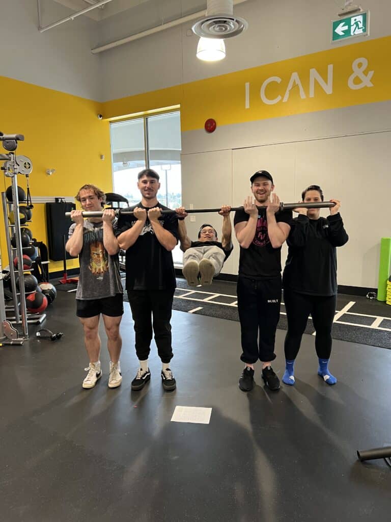 Trainers taking group photo together holding barbell with person hanging from it.